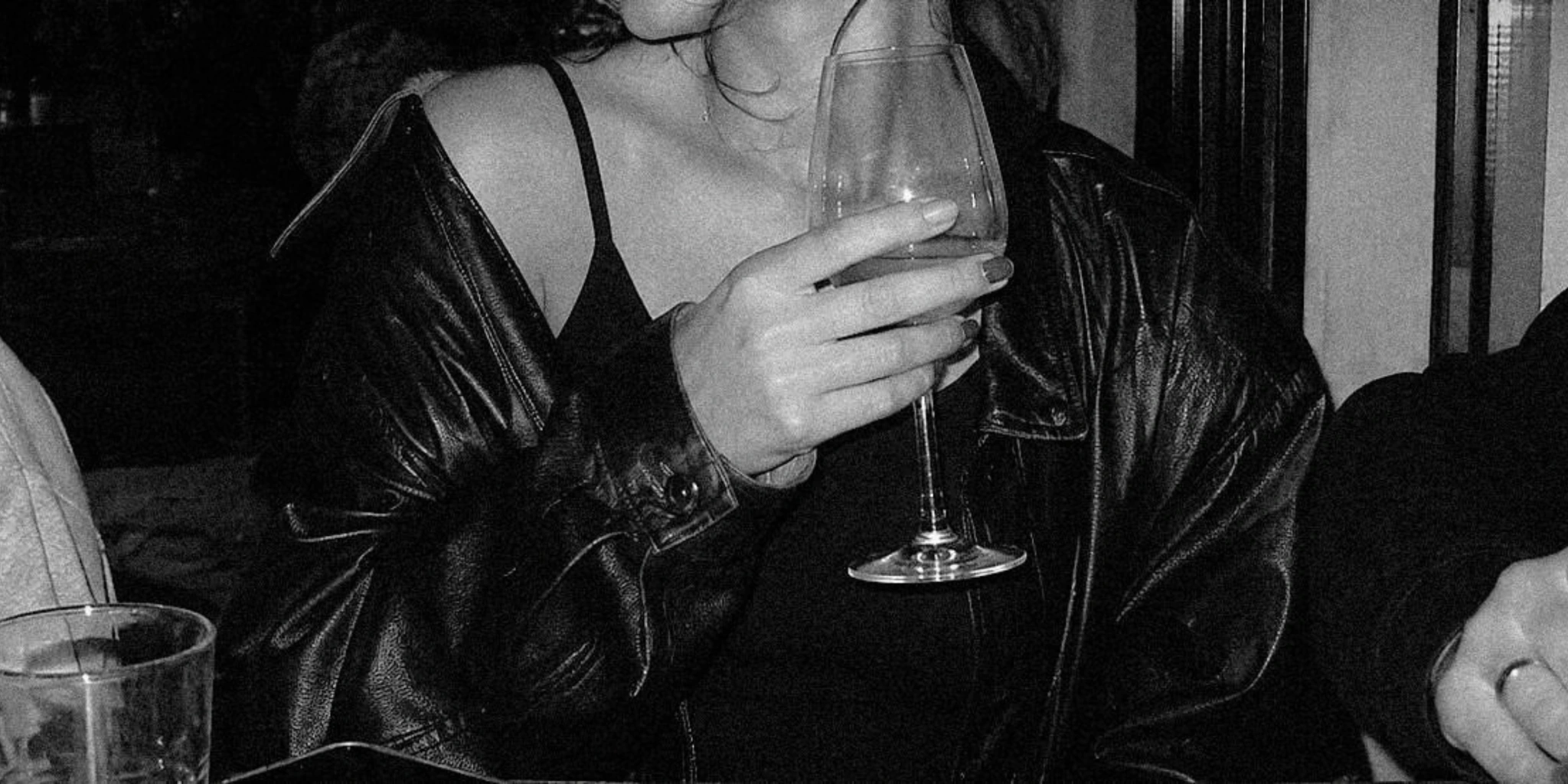 Black-and-white close-up of Maxine Meyer's hand holding a stemmed glass, with a black leather jacket
